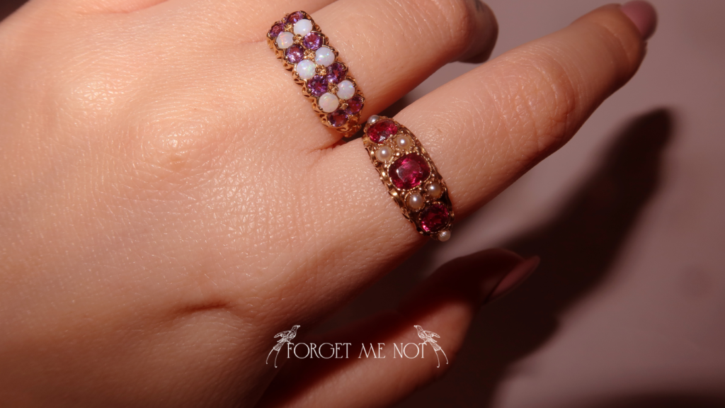 A close-up image of a hand wearing two ornate rings, each adorned with colorful gemstones and pearls. The lighting highlights the intricate details of the jewelry. Below the hand, the words "FORGET ME NOT" are elegantly written in delicate script, flanked by two small bird illustrations.